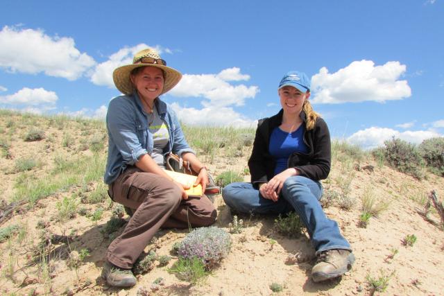 Two smiling young women sit on a sandy hill surrounded by sparse grasses and other vegetation.