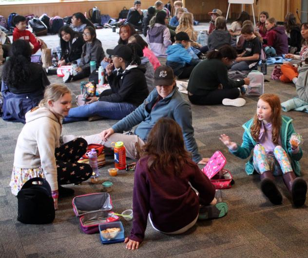 A large group of students sit on a carpeted floor and eat their packed lunches while having engaging conversations.