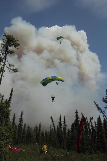 Two blue and yellow parachutes descend to the ground with a large smoke column in the background. 