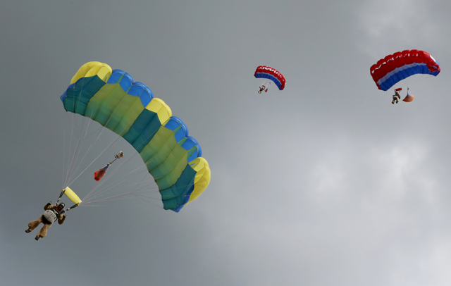 Three smokejumper descend underneath brightly colored parachutes set against a cloudy sky. 