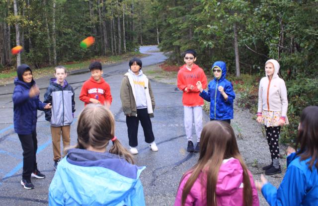 A group of young students stand in a circle outside and toss two Koosh balls to their partners.