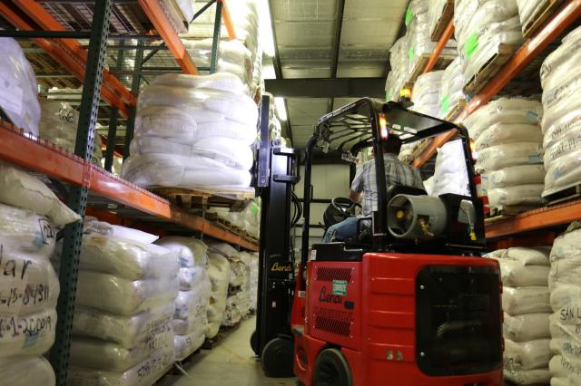 An orange forklift in front of tall pallet shelves lifts a pallet of white bags onto the shelf. The bags are filled with seeds.