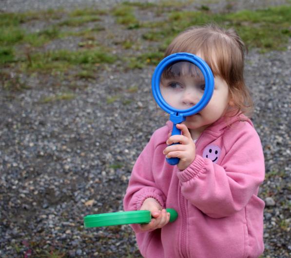A small toddler girl wearing all pink holds a blue plastic toy magnifying glass up to her eyes. Her other hand holds a green plastic magnifying glass.
