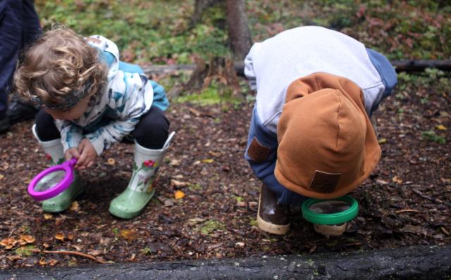 Two small children look through a magnifying glass on a nature trail outside.