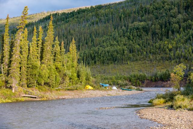 A camp with small inflatable boats is set up on the gravel bar along the forest lined river. 