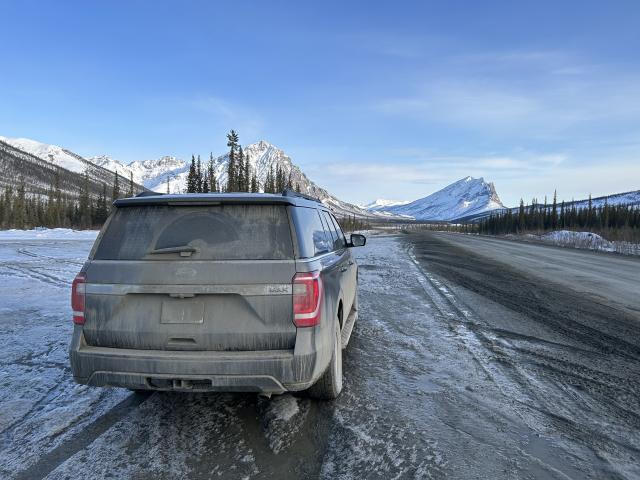 A very muddy vehicle parked alongside an icy road, with snowy mountains in distance