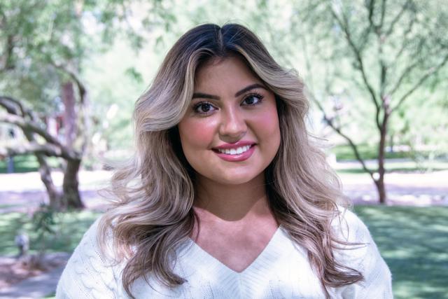 A woman wearing a white v-neck sweater smiles. Green trees are in the background.