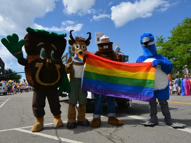 Four mascots holding a pride flag with blue sky and white clouds above.