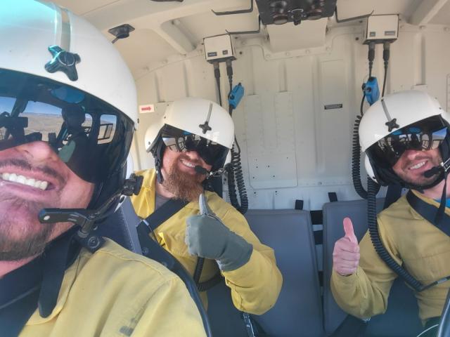 Three wildland firefighters in a helicopter smiling and giving thumbs up to the camera.