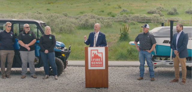 Utah's Governor Spencer Cox stands speaking behind a podium with Fire Sense members behind him.