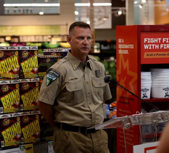 BLM Utah State Fire Management Officer Chris Delaney stands behind a podium addressing a crowd. Behind him is a shelf of fireworks and a shelf of gallons of water and buckets for dousing campfires and fireworks.