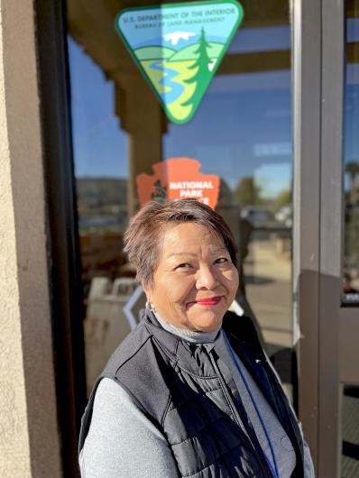 Gloria Benson, BLM Arizona Strip Distrcit Tribal Liaison, smiles in front of a window with the BLM and National Park Service logos.