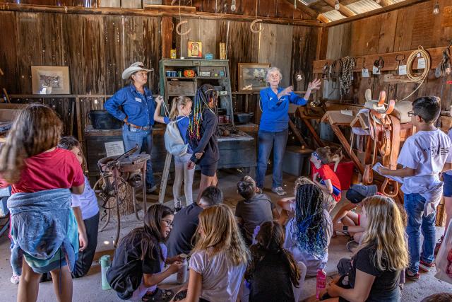 Group of students sitting and standing in an old barn filled with ranching equipment. Two adults are standing in front of the students.
