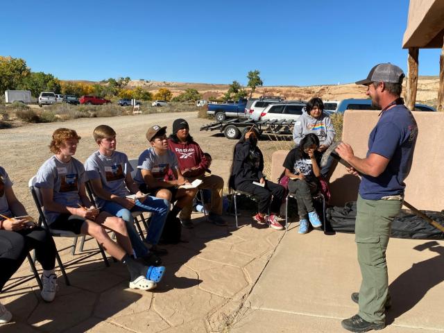 Students sit on folding chairs outside listening to a firefighter talk at Natural Resources Day event.