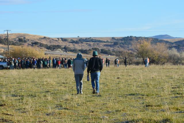 Two people walking in an open pasture field toward a large, gathered crowd. 