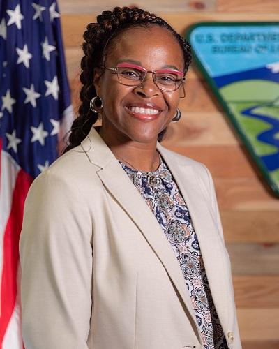 person with dark hair and glasses wearing a collared shirt posing in front of the American flag