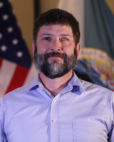 person with short hair and beard wearing a collared shirt posing in front of the American flag