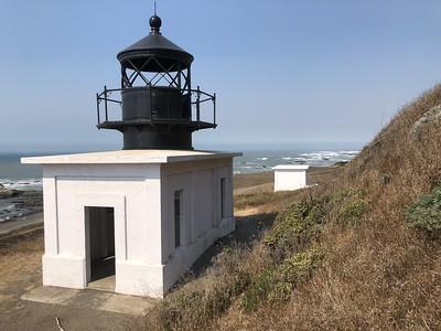 A white light house and fog signal building on the coast.