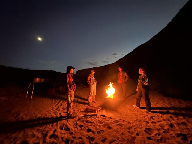 Members of the team huddle around a fire on a riverside beach with the dark sky silhouetting mountains behind them as the moon can be seen in the far left.