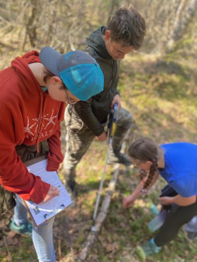 One students records data while two other students kneel and inspect a fallen log. 