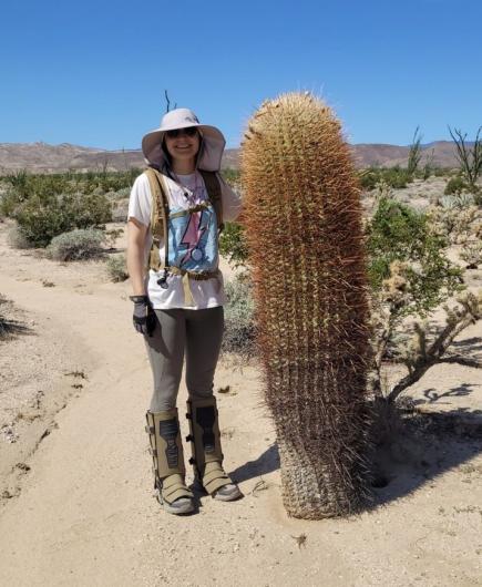 A young woman, smiling, wearing a sun hat and snake gaiters, stands next a large cactus that is the same height as her. She is surrounded by green shrubs with a gray mountain in the background. 