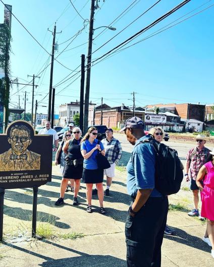 Individuals standing near a sign that reads “In memory of The Reverend James J. Reeb, American Unitarian Universalist Minister."