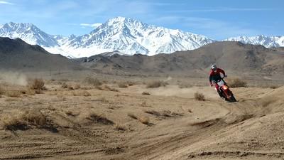 A dirt boke descends a hill with snow capped  mountains in the  background.