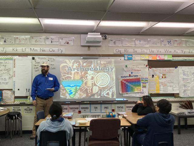 A photo of a man standing in front of students in an elementary school classroom, with display behind him that says "Archaeology."