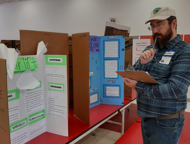 A photo of a man standing next to a row of table top displays containing various science projects. He is holding a pencil and looking at the displays with a clipboard. 