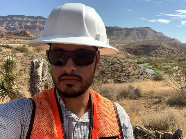 Carlos Joya, BLM Arizona's state engineer, takes a selfie. He is wearing a white hardhat, black glasses, and an orange high-visibility vest. The Virgin River Campground area is behind him.