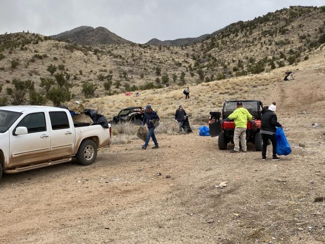 Trucks and off-road vehicles are positioned to help volunteers on the desert hillsides. People are seen loading full trash bags into the backs of these vehicles. Others are on the landscape in the background picking up trash