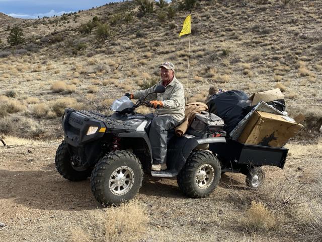 Man on 4-wheeler hauling a small trailer filled with garbage on a dirt road.