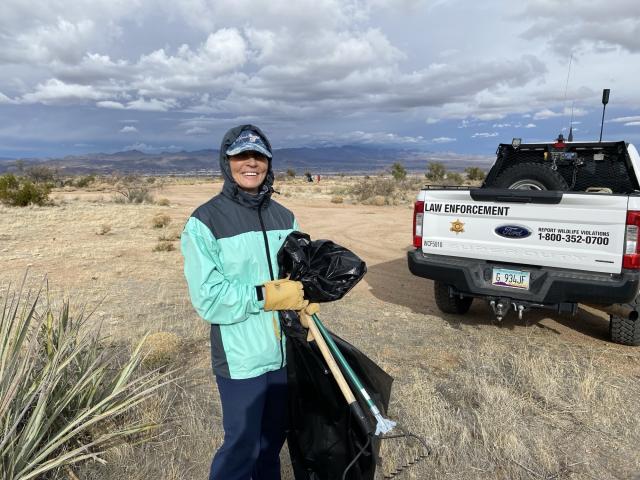 Desert clean-up volunteer stands clutching a black garbage bag and trash grabbers . The tailgate of an Arizona Game and Fish vehicle is visible to the right in the background. 
