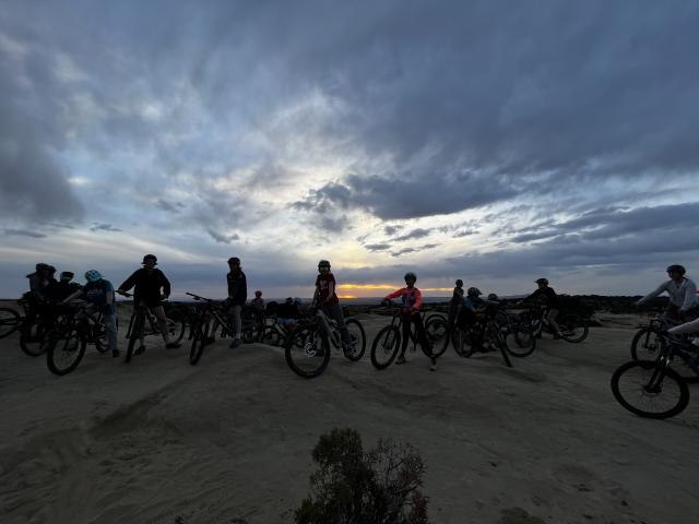 A photo of a group of youth mountain bike riders sitting atop their bikes on a desert trail, with an evening cloudy sky in the background.