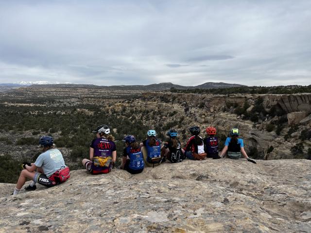 A photo of a group of youth mountain bike riders sitting on a ledge, with an evening cloudy sky in the background.