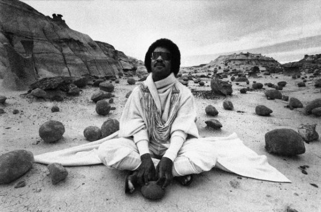 Alt Text: Photo shows black and white image of Stevie Wonder sitting on the ground touching a round rock.