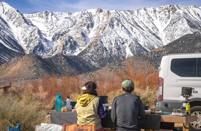 A couple at  a picnic table eats a meal while looking at a snowy  mountain.