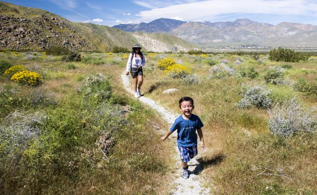 A child runs down a gravel trail in front of an adult. The trail is surrounded by short vegetation with mountains rising in the background.