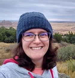 smiling person wearing glasses and a beanie standing in a meadow