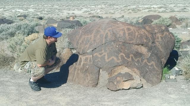 The photo shows a desert landscape with a man in a blue ball cap and sage green clothing, squatting down next to a large boulder that is covered in petroglyphs.
