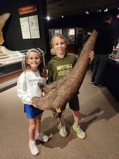 Two young children hold a triceratops horn cast that is almost as big as they are.