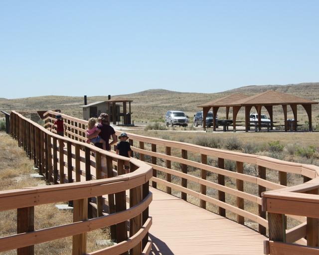 Photo shows a wooden boardwalk with rails on each side. Bathrooms, a covered picnic area, and a parking lot are in the middle ground. Arid hills are in the background. A woman, holding a young child on her hip, is walking away from the camera with one child in front of her and another by her side.