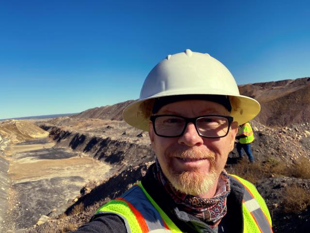 A man wearing a white hard hat and safety vest takes a selfie.