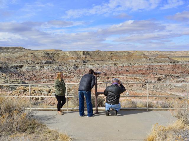 Three people look out at an expanse of colorful badlands. One is looking through permanent binoculars on a stand.