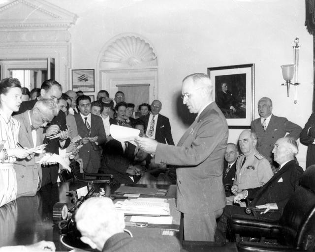 Image shows President Harry S. Truman standing at a desk and announcing the surrender of Japan to a group of reporters.  