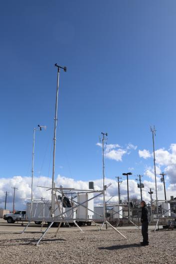 A person stands looking up at 30-foot metal poles with weather vanes on the top supported by tripod legs.
