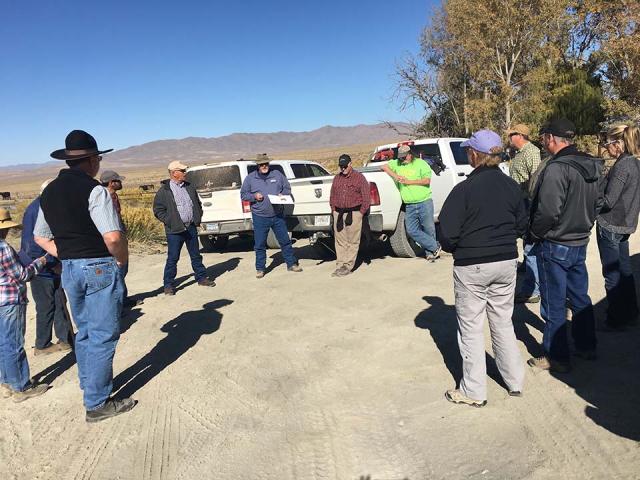 A group of adults in field clothes meet in a parking lot surrounded by blue skies and trees.