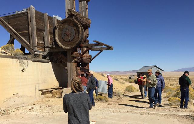 Surrounded by desert and a blue sky, a group of adults investigate a large piece of historic mining equipment in an old ghost town.