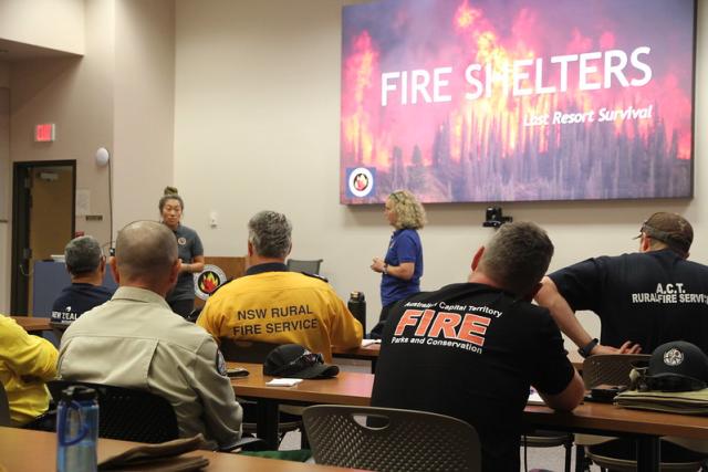 People in fire shirts sit and listen to a presenter indoors next to a projection screen featuring the words "fire shelters."