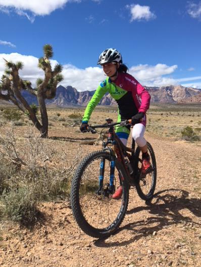 A smiling rider in helmet and longsleeve shirt rides a mountain bike across desert flagged by sagebrush and joshue trees